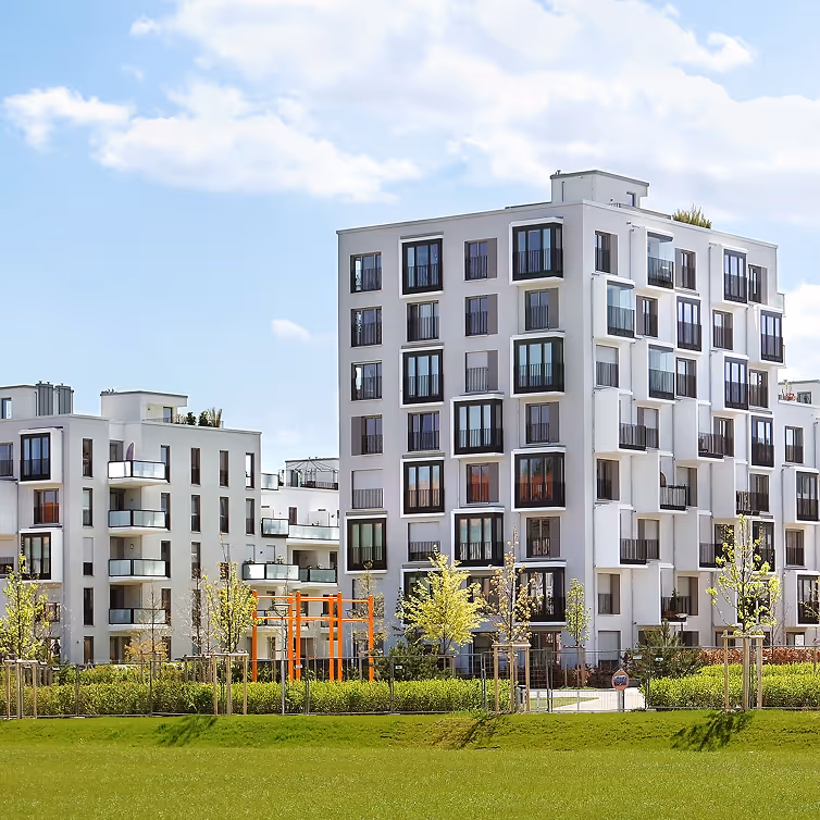 Modern white apartment buildings with large windows and balconies, surrounded by young trees and green lawn under a partly cloudy sky.