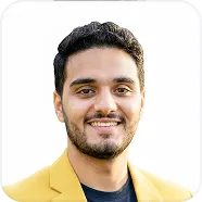 Smiling man with dark curly hair and beard wearing a yellow jacket over a dark shirt against a white background.