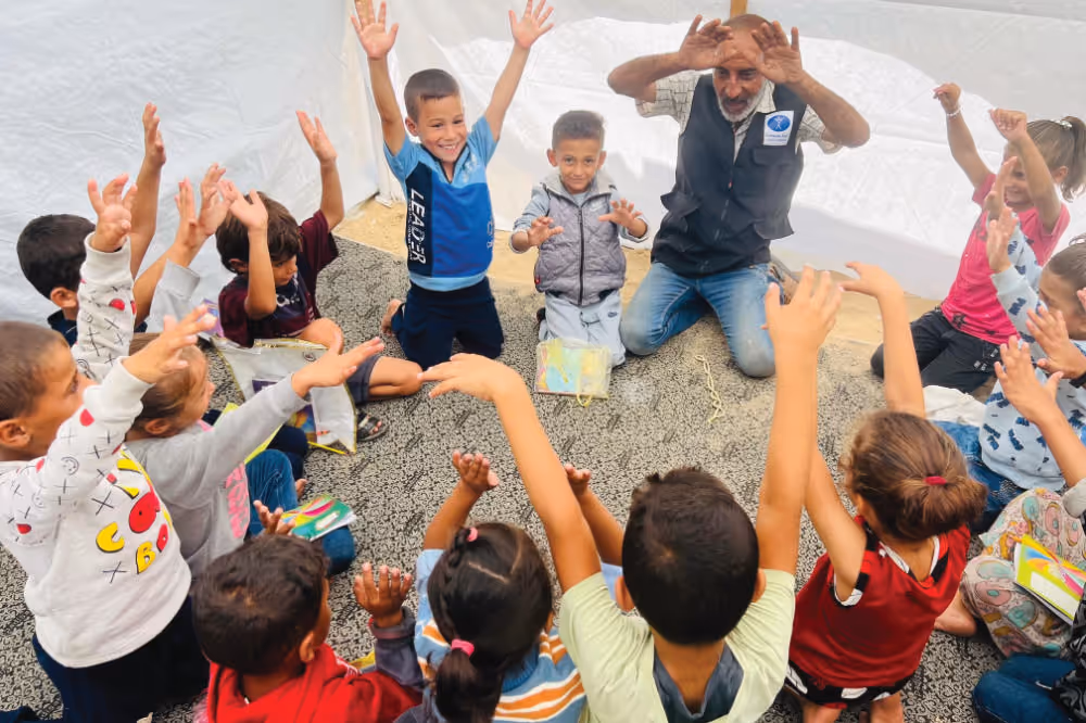 A group of children sitting in a circle on a patterned carpet, raising their hands and smiling while a man kneels among them with hands raised, inside a tent.