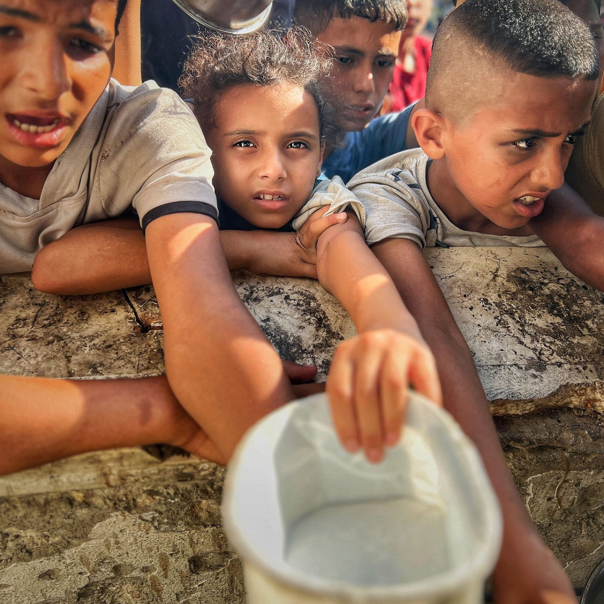 Several children reaching out with outstretched arms holding an empty container, expressing urgency or need.