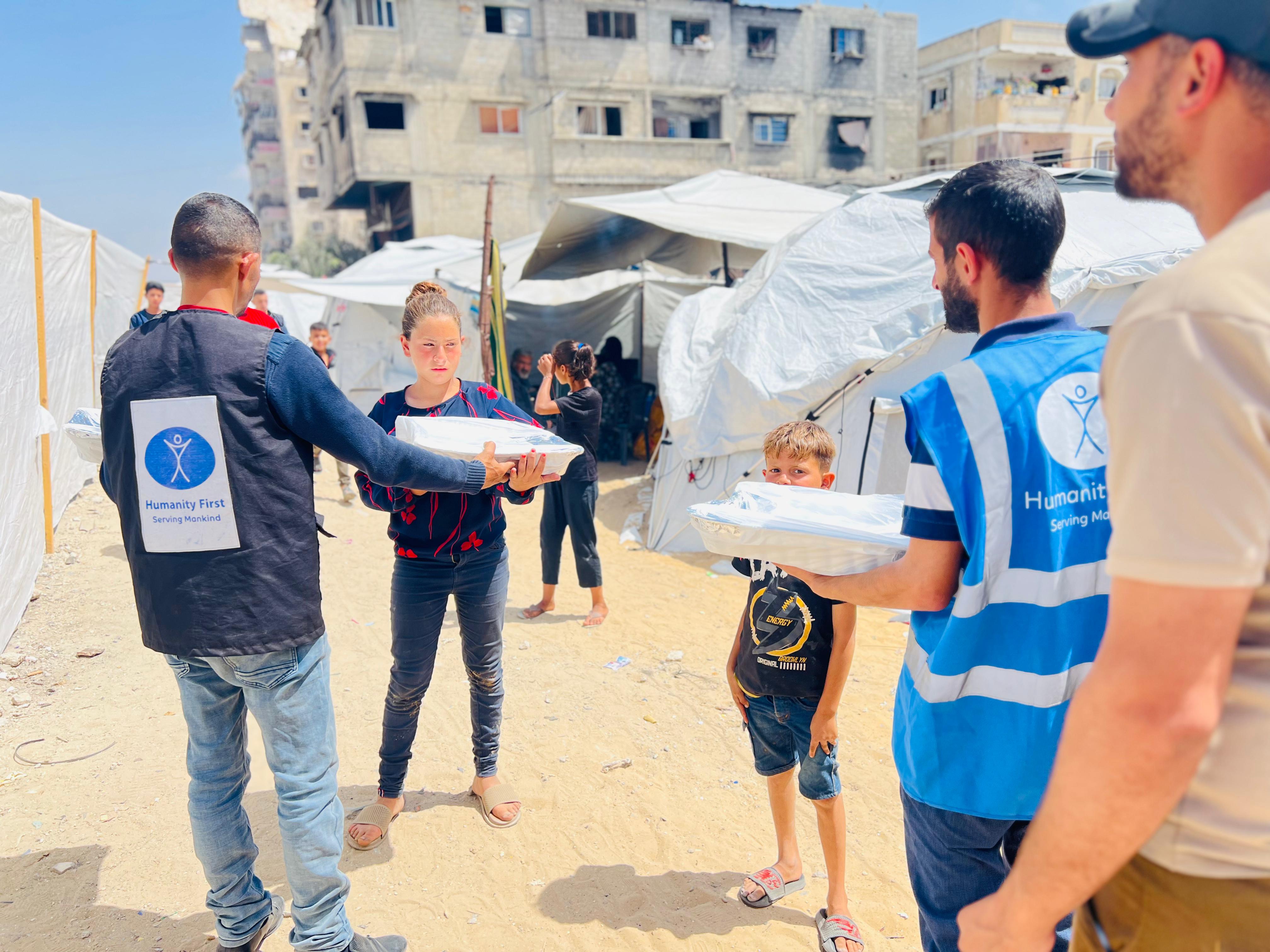 Volunteers from Humanity First distributing food packages to children in a tented refugee camp with damaged buildings in the background.