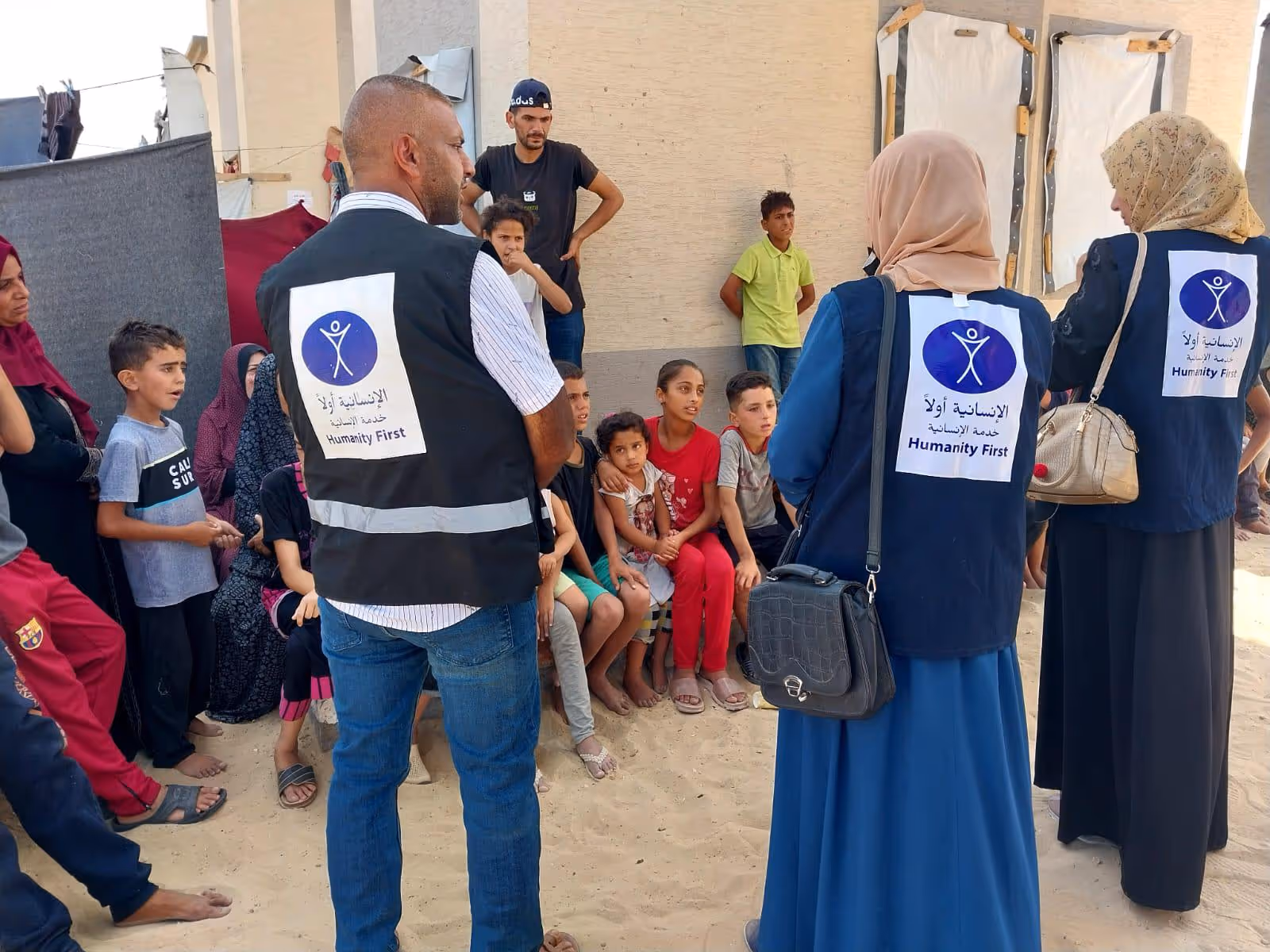 Three Humanity First workers wearing vests talk to a seated group of children and adults in a sandy outdoor setting.