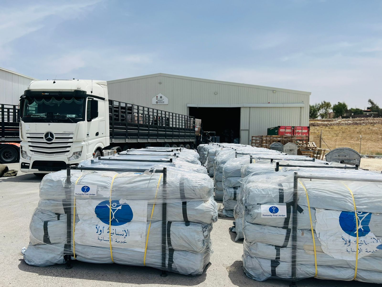 Stacks of packaged humanitarian aid tents labeled Humanity First Jordan next to a white Mercedes truck outside a warehouse.