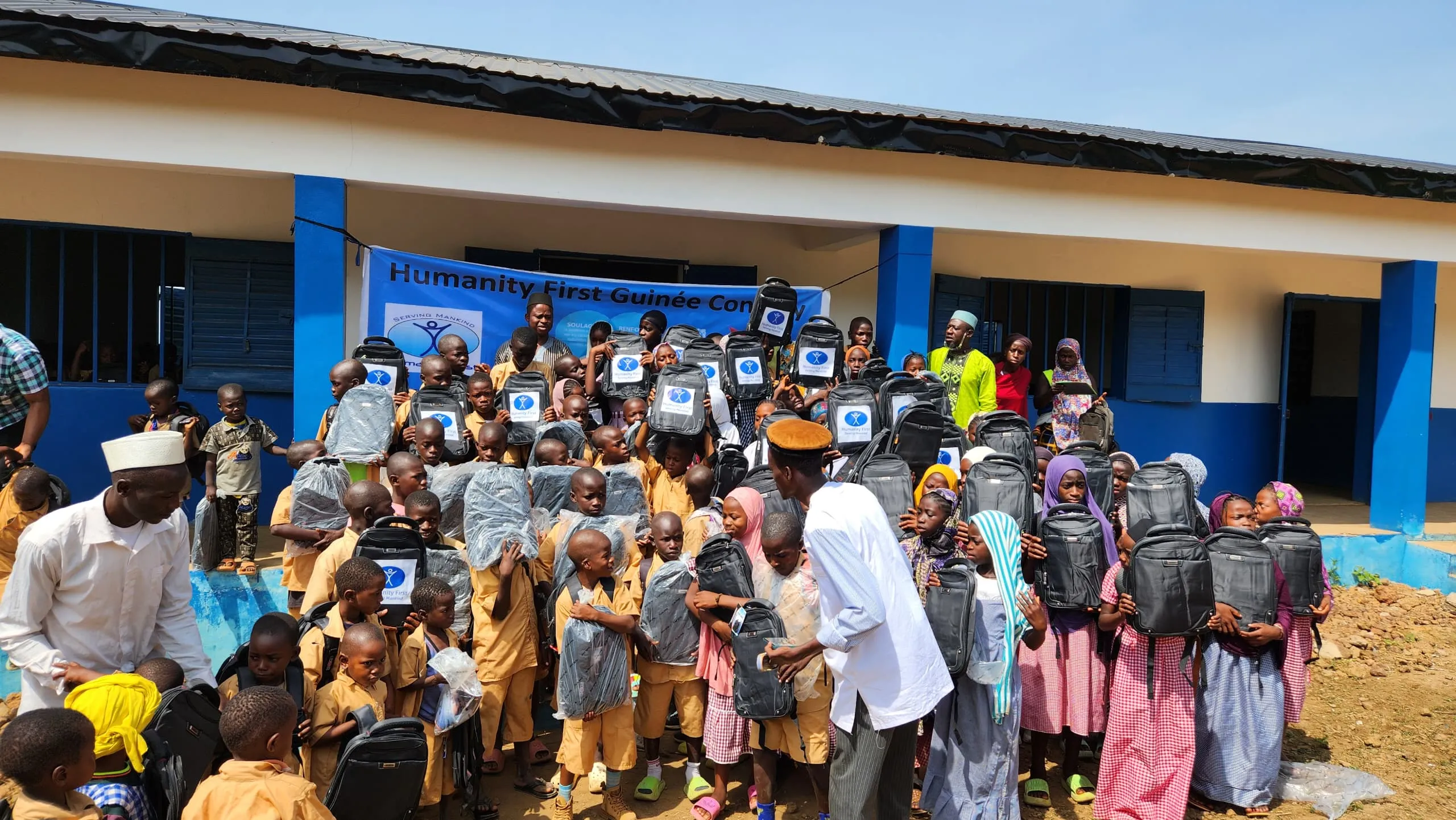 Group of children and adults outside a blue and white building, many holding new black backpacks with Humanity First logos.
