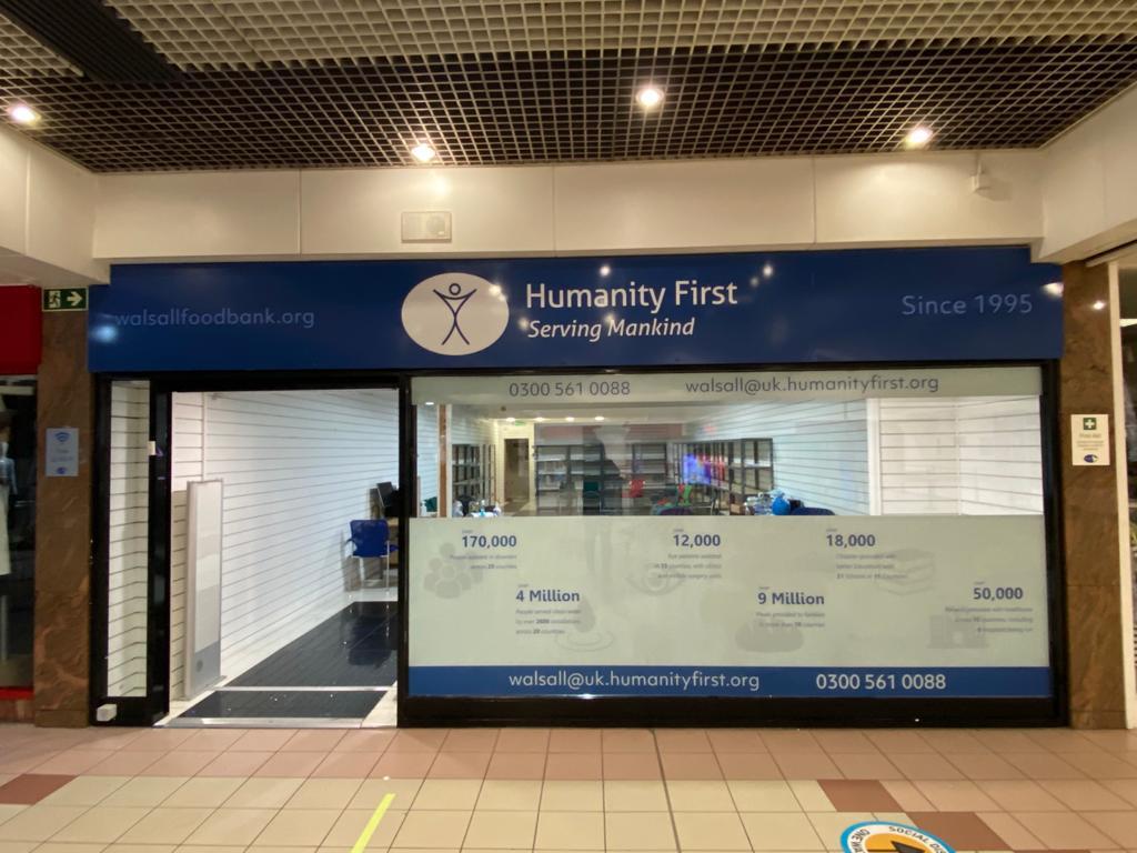 Front view of Humanity First Walsall food bank with blue signage displaying contact details and impact statistics on the glass window.