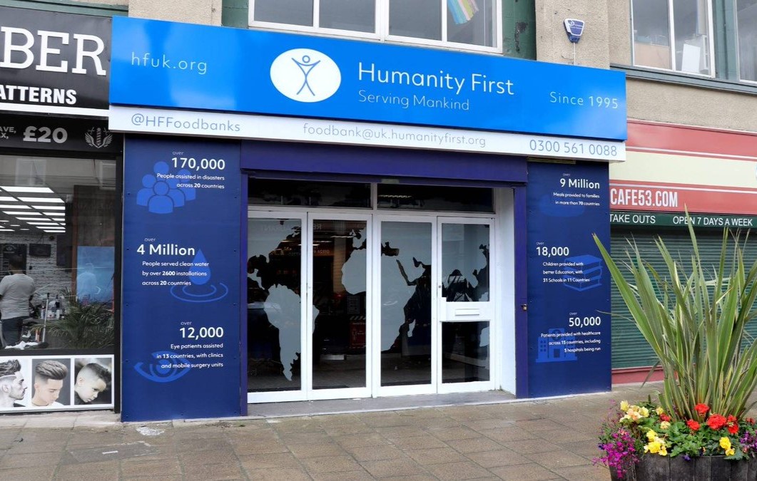 Front of Humanity First food bank with large blue signage displaying statistics about people assisted, meals provided, and patients served globally.