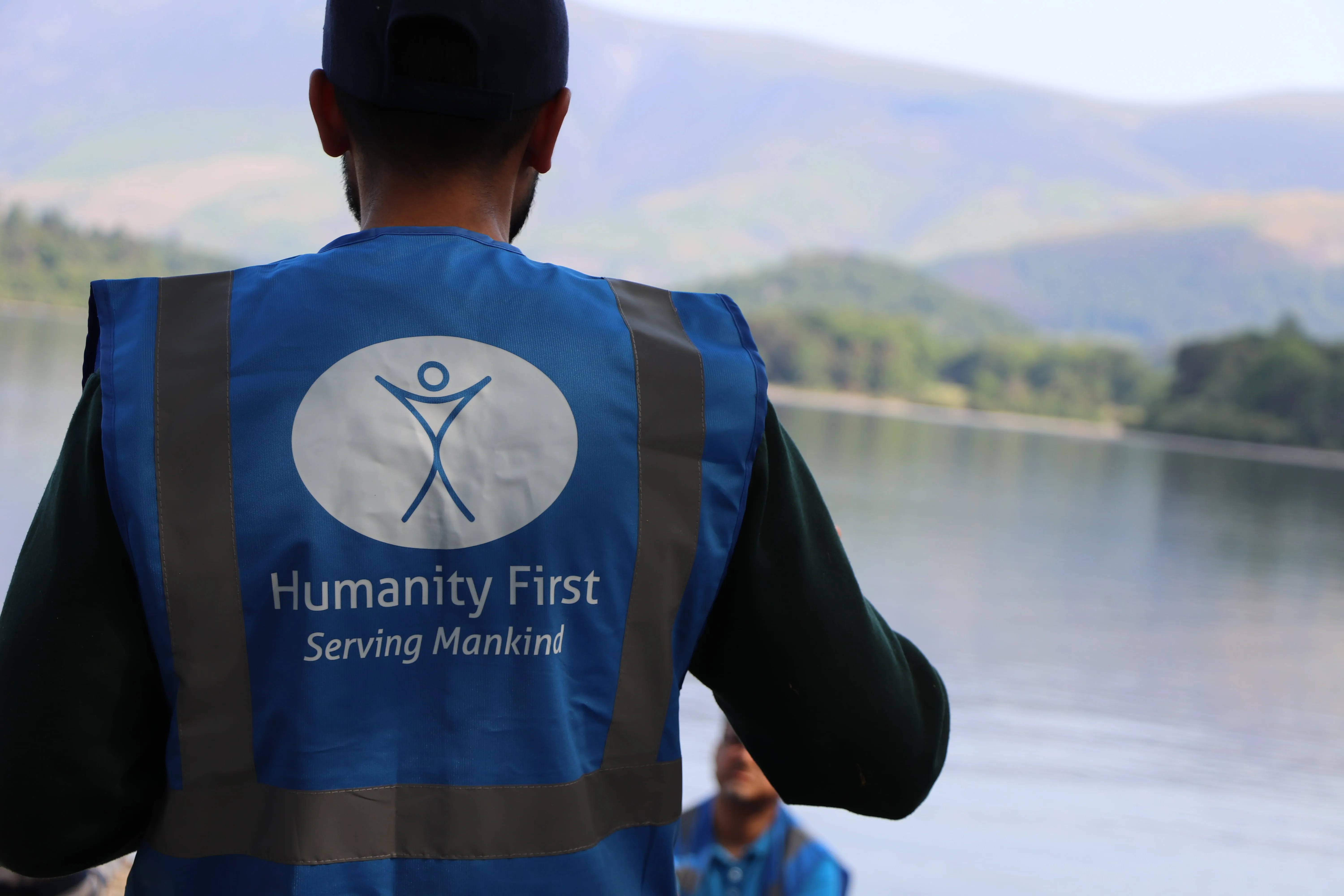 Person wearing a blue Humanity First vest overlooking a calm lake with mountains in the background.