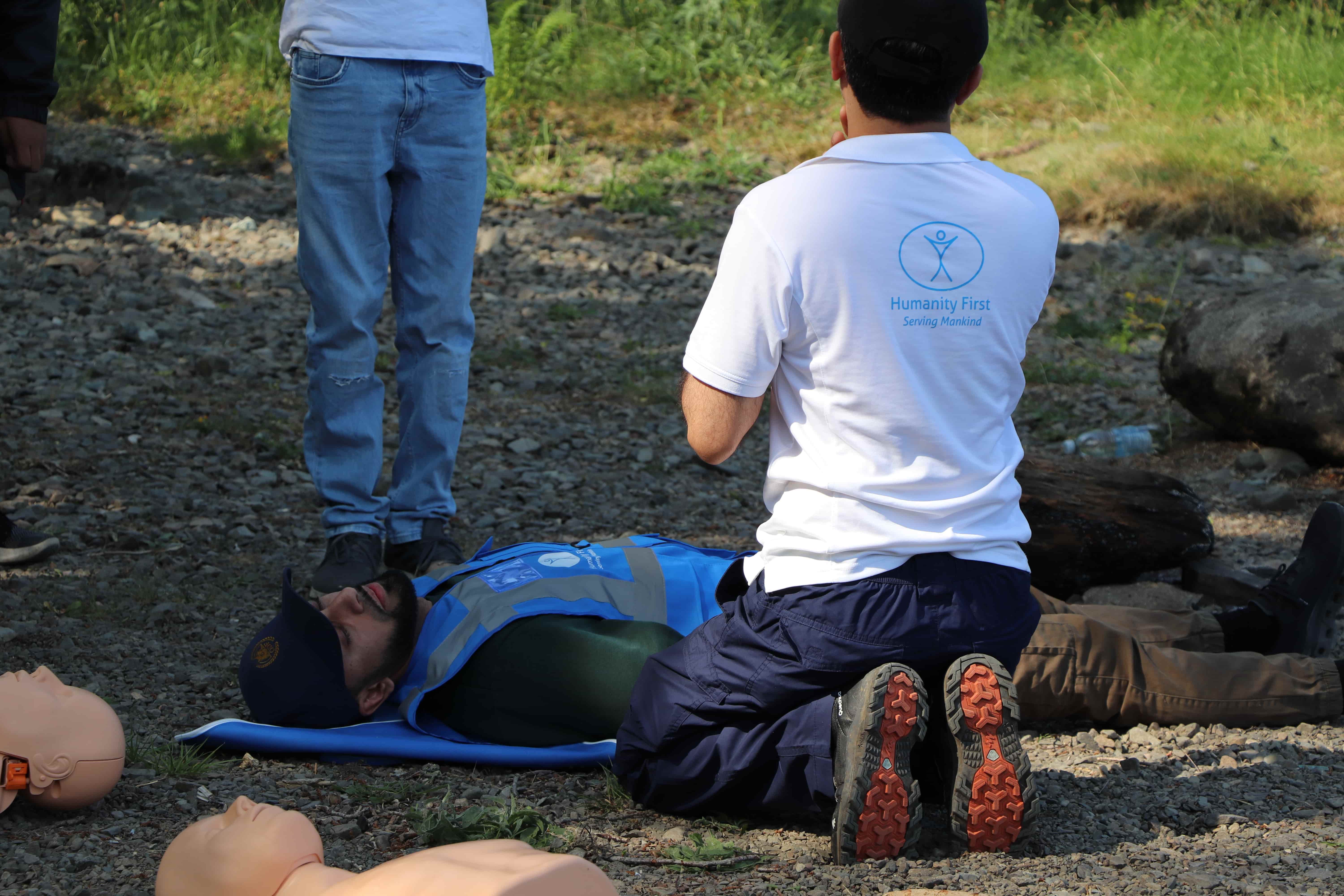 Man in a white shirt kneeling on rocky ground performing CPR on a person lying on a blue mat, with CPR training mannequins nearby.