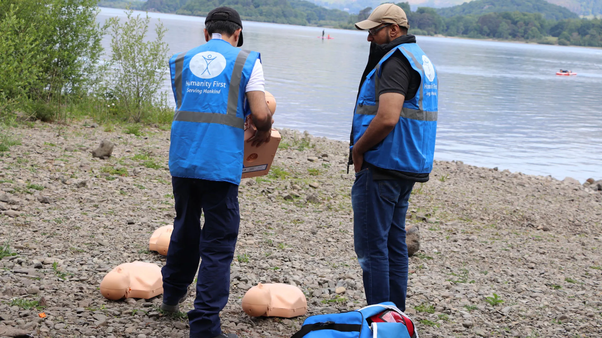 Two men wearing blue Humanity First vests conducting CPR training with mannequins on a rocky lakeside shore.