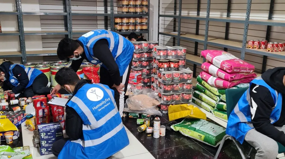 Volunteers in blue Humanity First vests sorting and organizing canned goods and food supplies in a storage room with empty shelves.