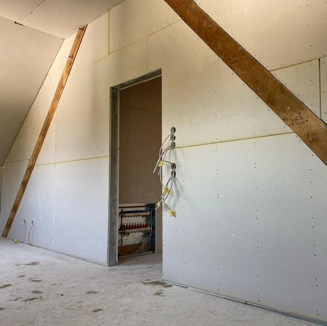 Weißes Waschbecken mit verchromtem Wasserhahn, goldfarbenem Seifenspender und grüner Pflanze vor schwarzen Wandfliesen.