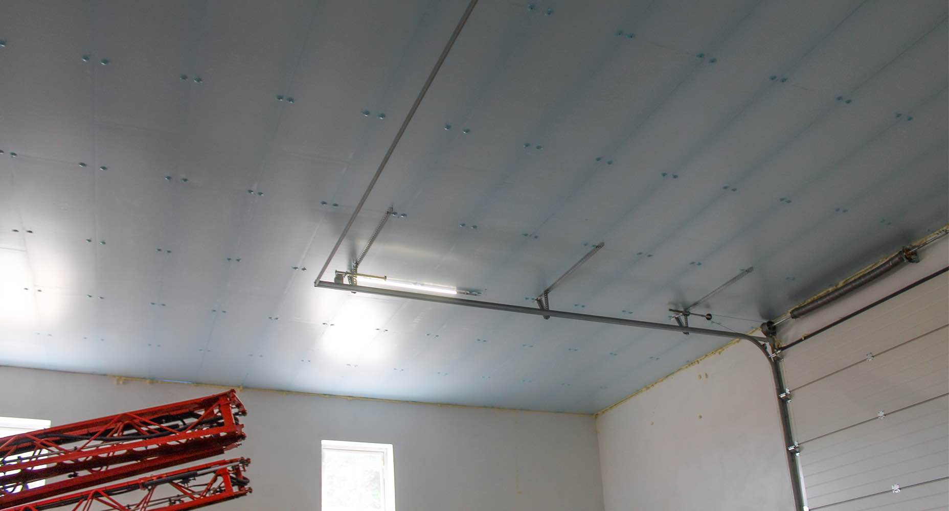 Wnętrze garażu z błękitnymi panelami izolacyjnymi XPS Agro na suficie, prowadnicami i bramą segmentową oraz czerwoną maszyną rolniczą do oprysków stojącą przy ścianie z oknami.