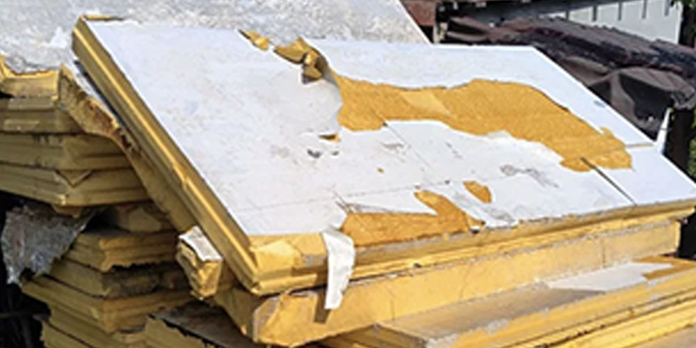 Uszkodzona płyta PIR z odchodzącą białą naklejaną folią aluminiową (50 µm). Widoczne odwarstwianie okładziny i odsłonięcie żółtego rdzenia – klasyczny przykład niższej trwałości klejonej folii w porównaniu do integralnego naskórka wysokiej gęstości w panelach XPS Agro.