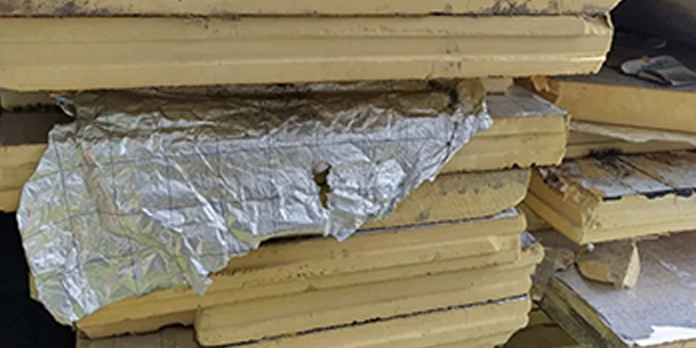 Stos żółtych płyt termoizolacyjnych PIR/PUR z rozerwaną, pomiętą i odchodzącą folią aluminiową (pazłotko / sreberko). Widoczne mechaniczne zniszczenie warstwy ochronnej – klasyczny przykład jednej z głównych wad popularnych materiałów termoizolacyjnych: łatwego rozwarstwiania i uszkodzeń folii aluminiowej podczas magazynowania, transportu, montażu lub użytkowania i mycia myjką ciśnieniową.