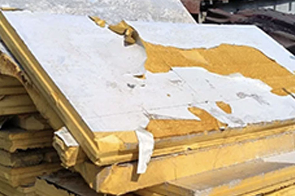 Uszkodzona płyta PIR z odchodzącą białą naklejaną folią aluminiową (50 µm). Widoczne odwarstwianie okładziny i odsłonięcie żółtego rdzenia – klasyczny przykład niższej trwałości klejonej folii w porównaniu do integralnego naskórka wysokiej gęstości w panelach XPS Agro.