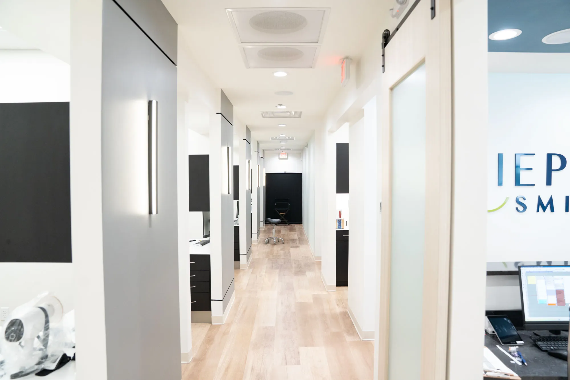Bright, modern dental or medical office hallway with light wood flooring, white walls, black fixtures, and a rolling stool at the far end.