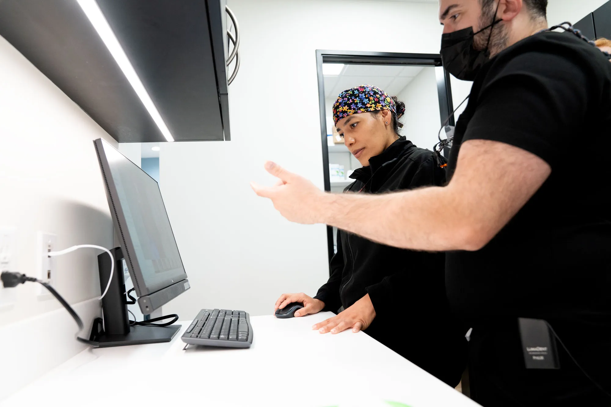 Two medical professionals in black scrubs review information on a desktop computer; the woman uses the mouse while the masked man gestures toward the screen.