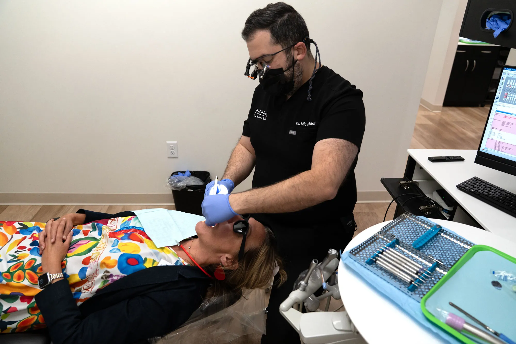 Dentist wearing black scrubs and magnifying glasses with a headlamp performs a dental procedure on a woman reclining in a dental chair, with dental tools and a computer monitor visible nearby.