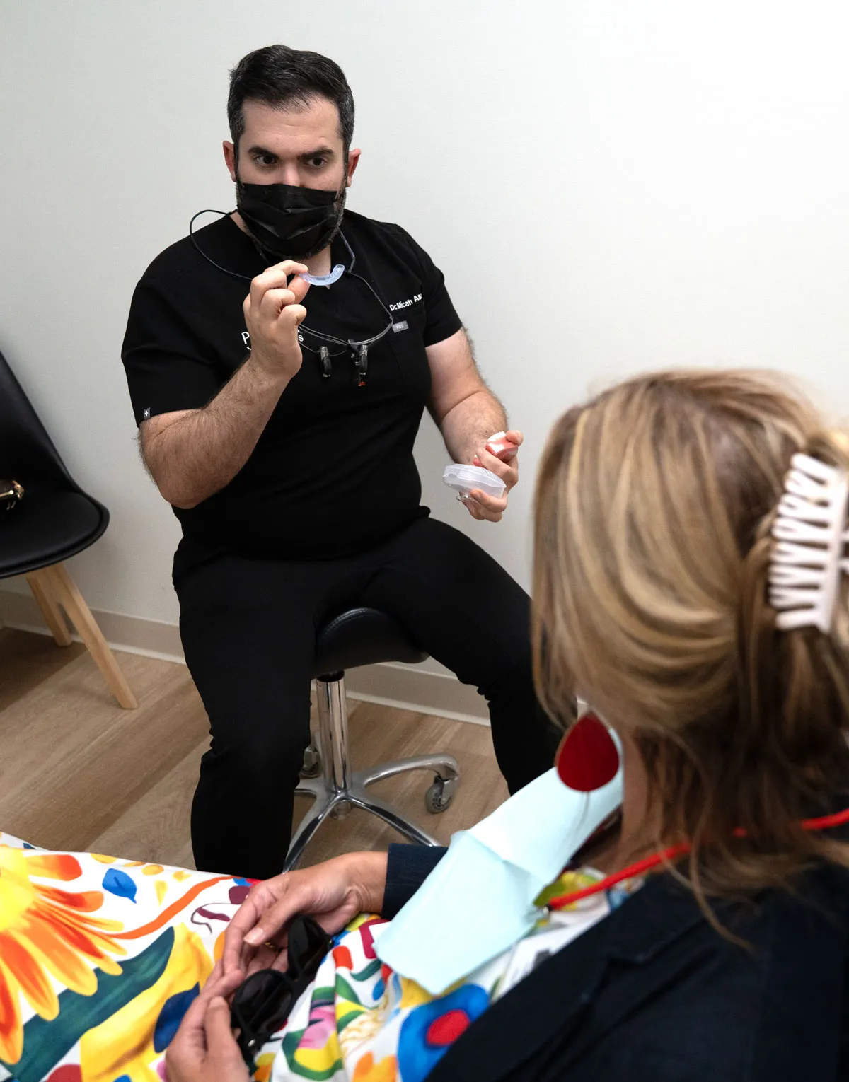 A dentist wearing black scrubs and a face mask holds a dental retainer and a container while explaining to a seated patient dressed in colorful clothing and holding sunglasses.