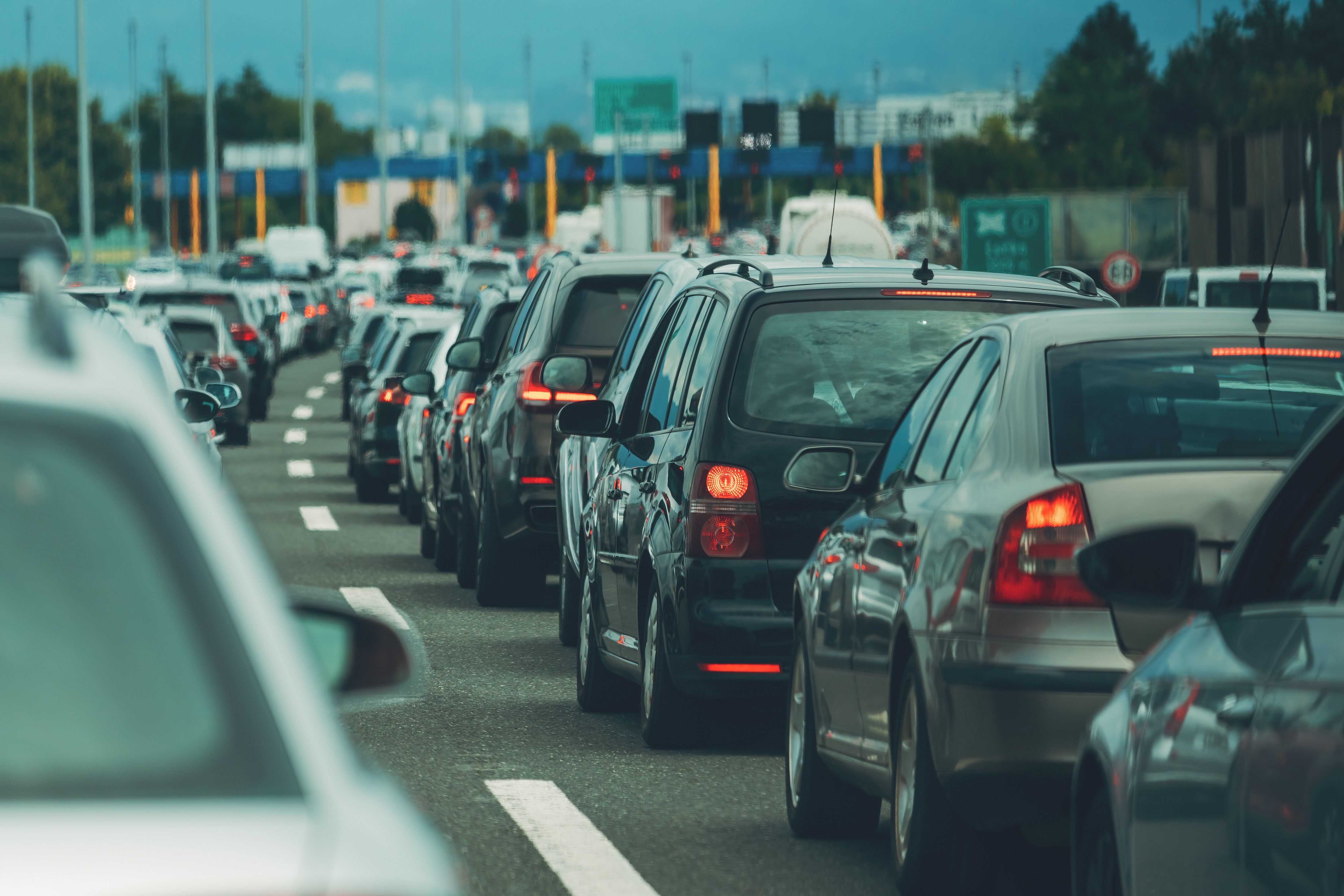 Long line of cars stopped in traffic on a multi-lane highway near a toll area, with brake lights illuminated and vehicles queued closely together.