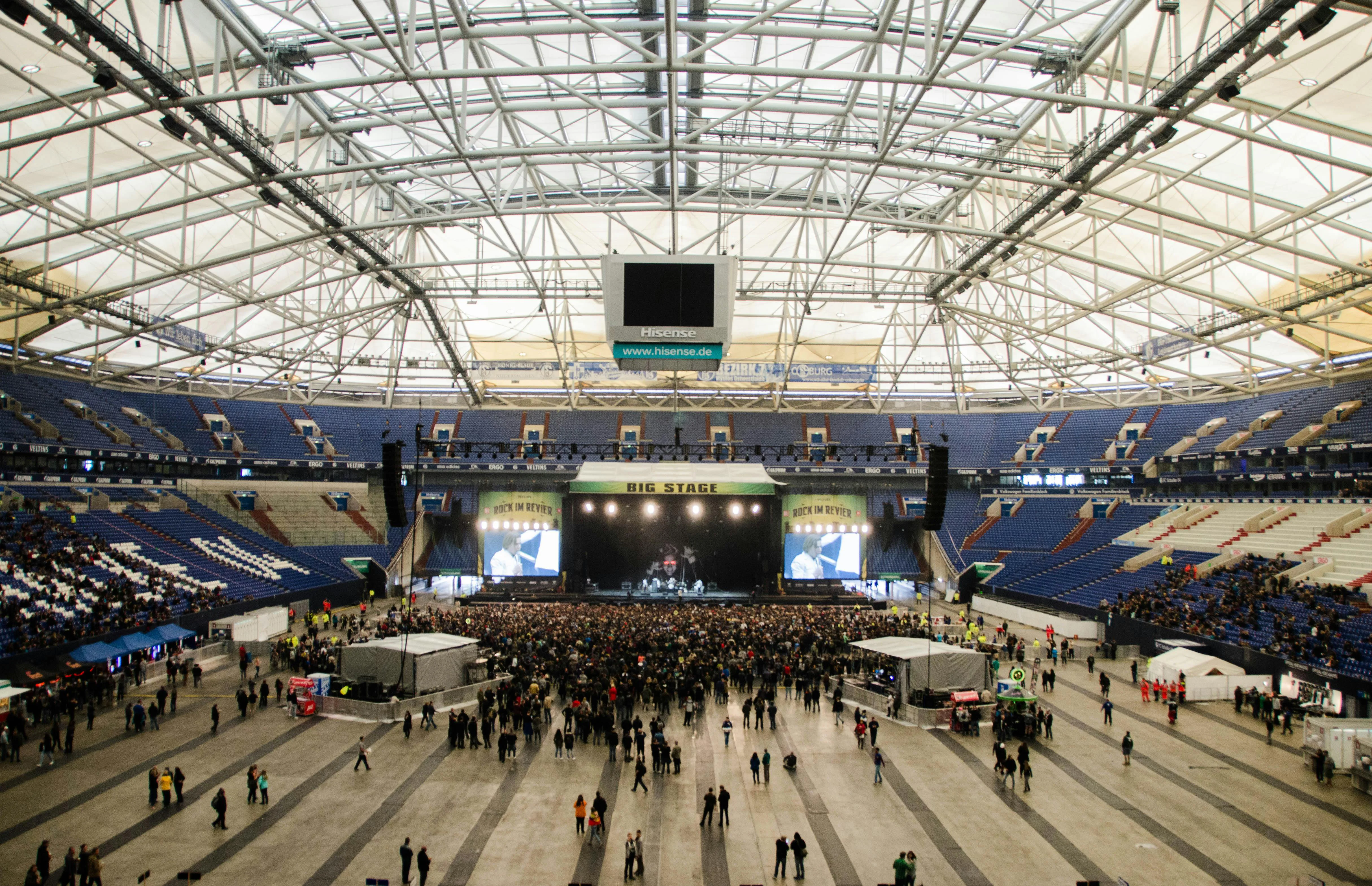 Wide view of a large indoor stadium with a crowd gathered in front of a central concert stage. The venue features a high steel-truss roof, tiered blue seating, large screens flanking the stage, and attendees spread across the arena floor.