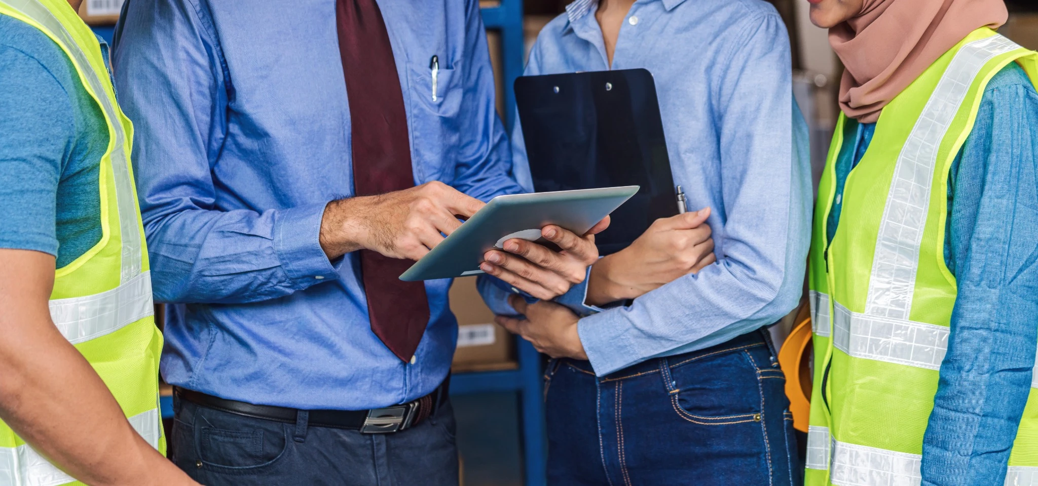 Close-up of a diverse team of warehouse or facility staff wearing high-visibility safety vests, reviewing information on a tablet and clipboard during an on-site operational meeting.