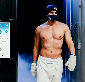 Shirtless man in mittens, towel, hat, mask, exiting whole body cryo therapy treatment.