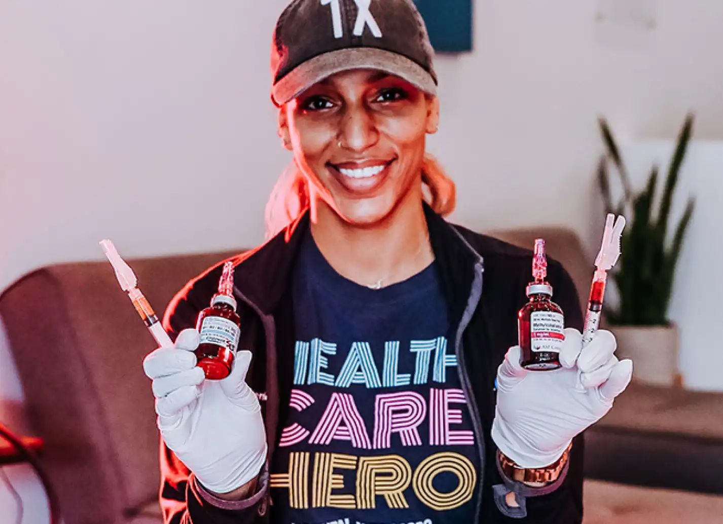 Female nurse wearing white rubber gloves and holding up one syringe and 1 vitamin vial in each hand.