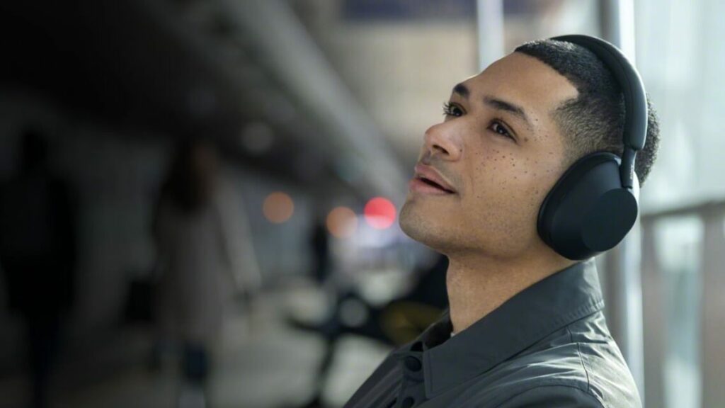 A young man with short, neatly trimmed hair and light freckles is wearing large, black over-ear headphones while leaning back slightly with a relaxed expression. He appears to be enjoying music or a podcast. The background is blurred, showing an indoor public space with people walking by and warm, ambient lights creating a soft bokeh effect.