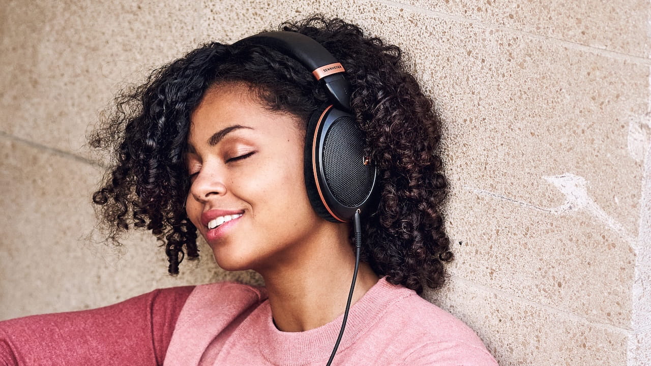 A young woman with curly hair and a warm smile is leaning against a textured beige wall with her eyes closed, wearing large, black and copper-colored over-ear headphones. She is dressed in a soft pink sweater and appears to be enjoying the music, immersed in a moment of relaxation.