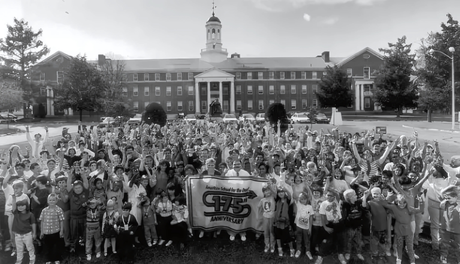 A large group of Deaf students, faculty, and community members gather in front of the American School for the Deaf to celebrate its 175th anniversary. The historic brick school building stands in the background, and people in the crowd cheer and wave, holding a banner that reads “175th Anniversary.”