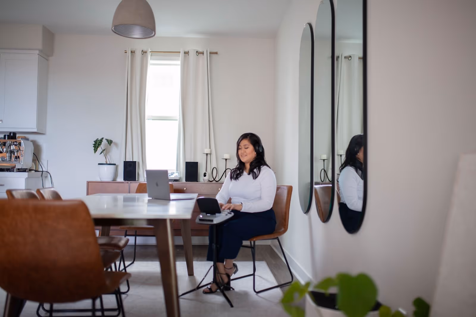 A woman sits at a dining table in a bright, modern apartment, wearing headphones and using a steno machine during a remote captioning session. A laptop sits in front of her, and the setting includes warm, minimalist decor.