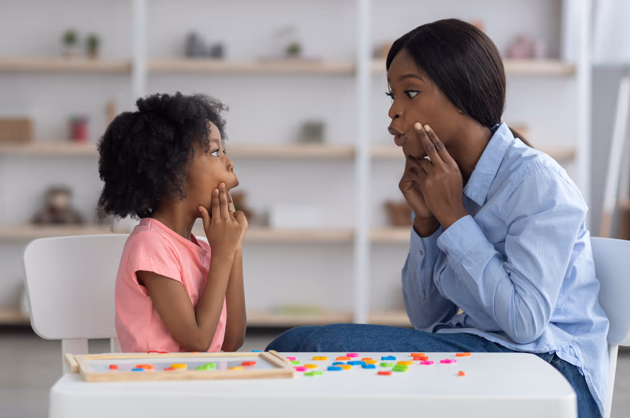 Speech therapist and young girl sitting across from each other at a table, both pointing to their mouths while practicing speech sounds during a therapy session with colorful letters scattered on the table.