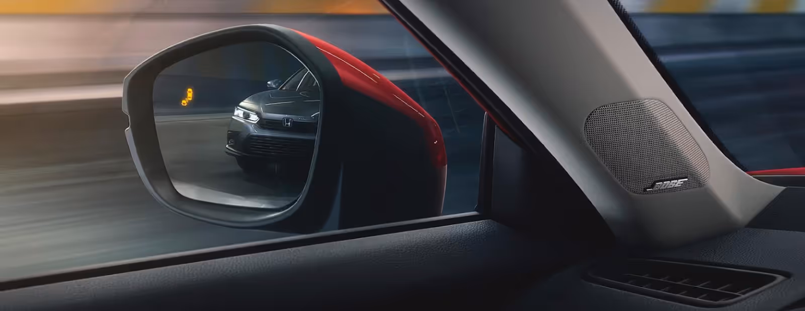 A side mirror view from inside a car shows a Honda approaching in the blind spot, with a yellow blind spot warning icon illuminated on the mirror. A Bose speaker is visible on the car’s interior pillar.
