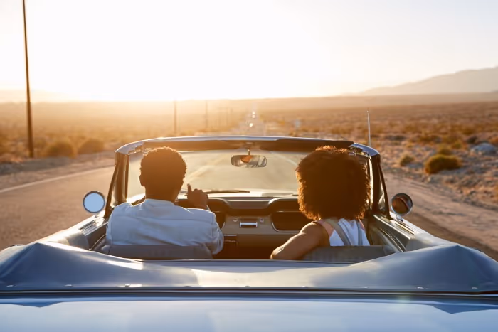 Two people drive a convertible down an empty desert highway at sunset, with clear skies and mountains in the distance.