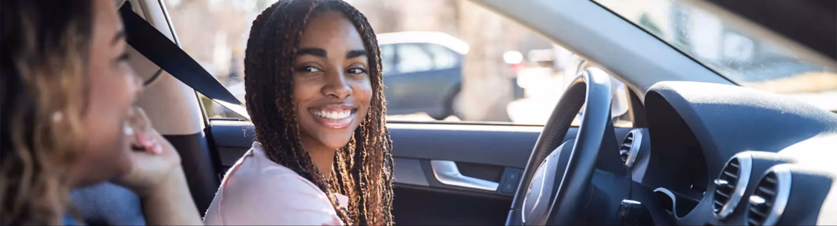A teenage girl smiles confidently while sitting in the driver’s seat, with another person sitting beside her in the passenger seat.