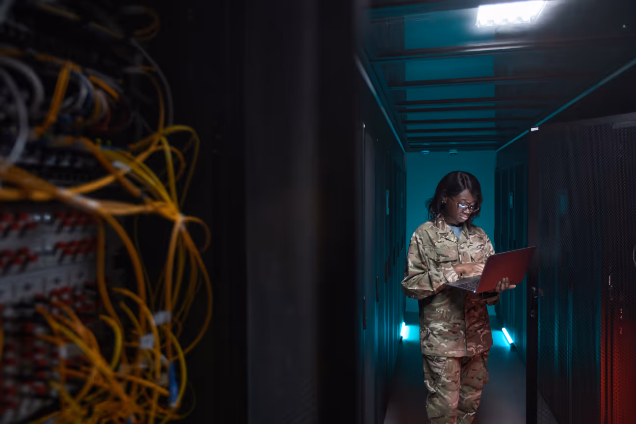 A U.S. military service member in camouflage uniform stands in a dark server room, working on a laptop. She is surrounded by computer servers and glowing lights, representing a tech-based military role.