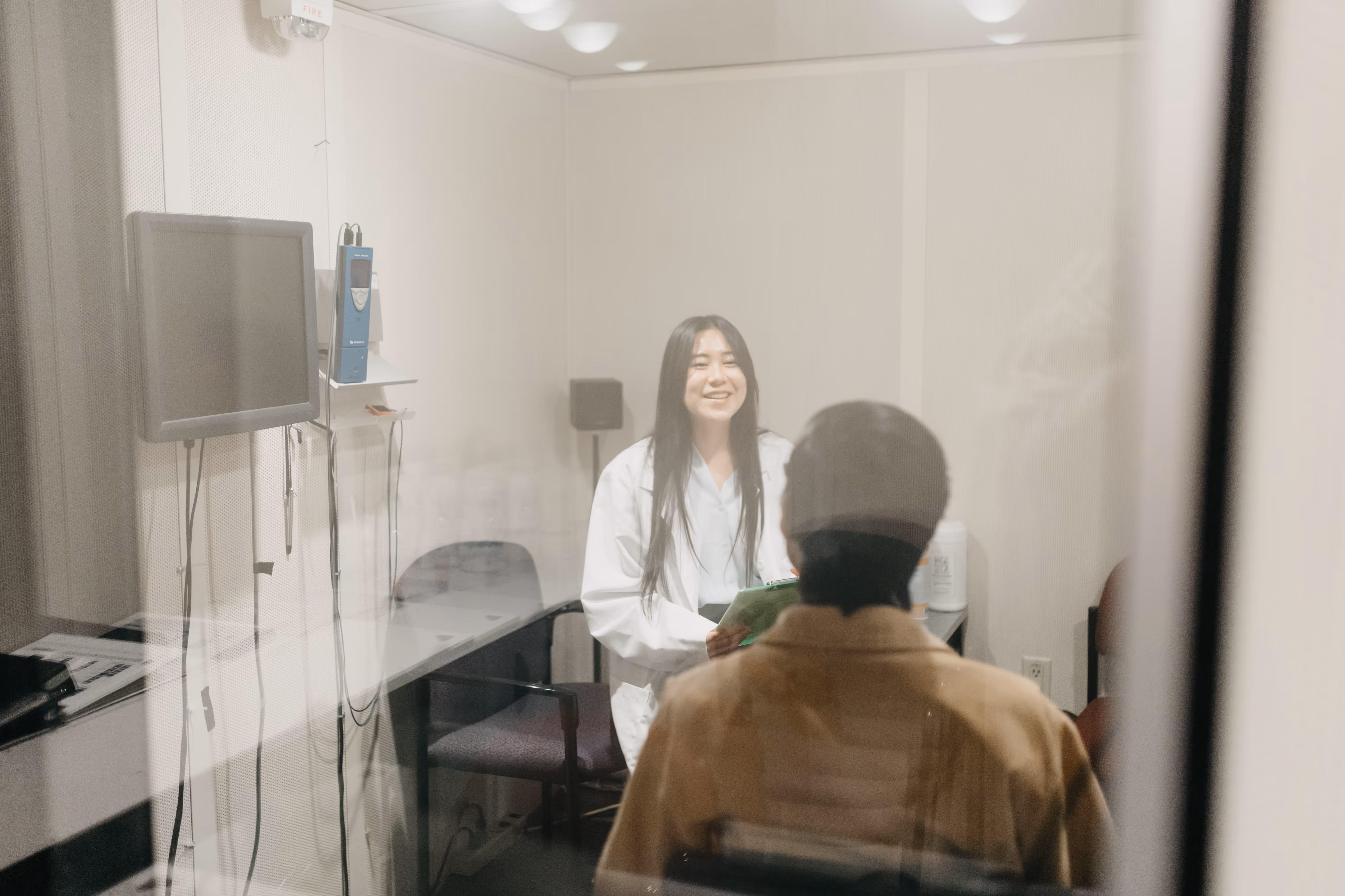 A view through a glass window into a soundproof audiology testing room. A female audiologist in a white coat smiles at a seated patient while holding a clipboard. Medical equipment and hearing test tools are visible in the background.