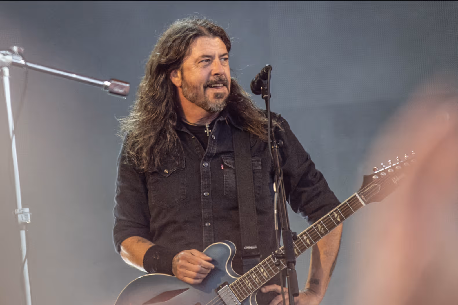 Dave Grohl of Foo Fighters stands on stage with a guitar, smiling mid-performance. He wears a black shirt with rolled sleeves and has long hair and a beard.