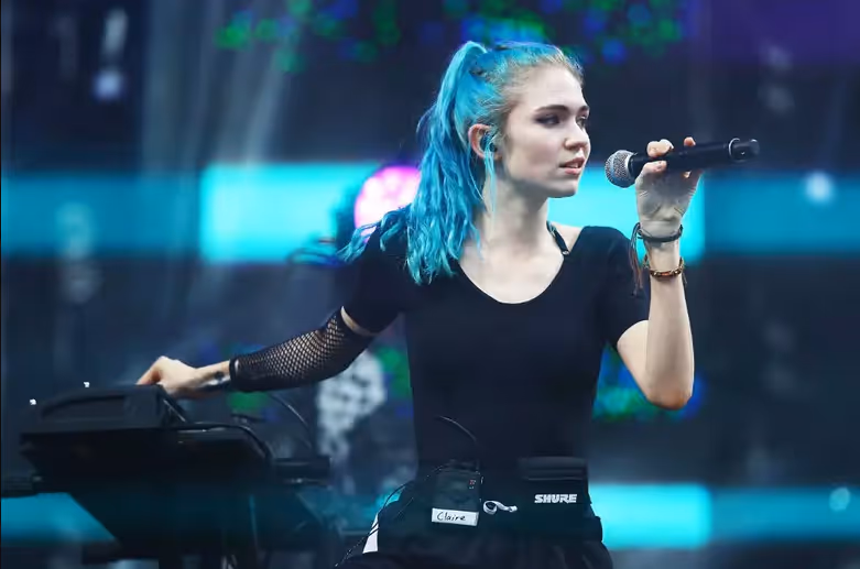 Grimes, with bright turquoise hair tied in a high ponytail, holds a microphone while performing on stage. She wears a black outfit with mesh sleeves and stands beside a keyboard under colorful concert lights.