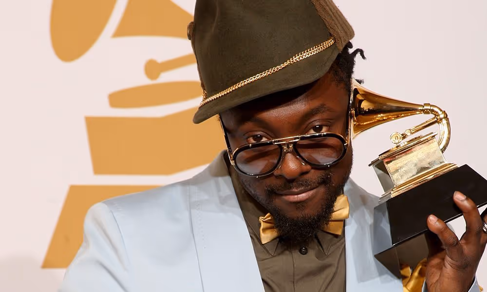 Will.i.am poses with a Grammy Award trophy, holding it up to his ear with a playful expression. He wears oversized glasses, a green cap with gold trim, a gold bow tie, and a light blue blazer. A Grammy logo is visible in the background.