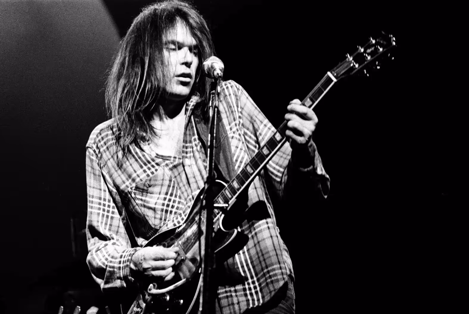 Neil Young performs live in a plaid shirt, strumming an electric guitar and singing into a microphone. The image is black and white with dramatic stage lighting.