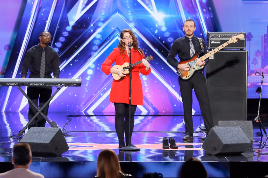 Mandy Harvey performs on the America’s Got Talent stage, wearing a bright red dress and standing barefoot while playing a ukulele. Two musicians—a keyboardist and bassist—stand behind her, and the AGT logo and lights form a vibrant backdrop.