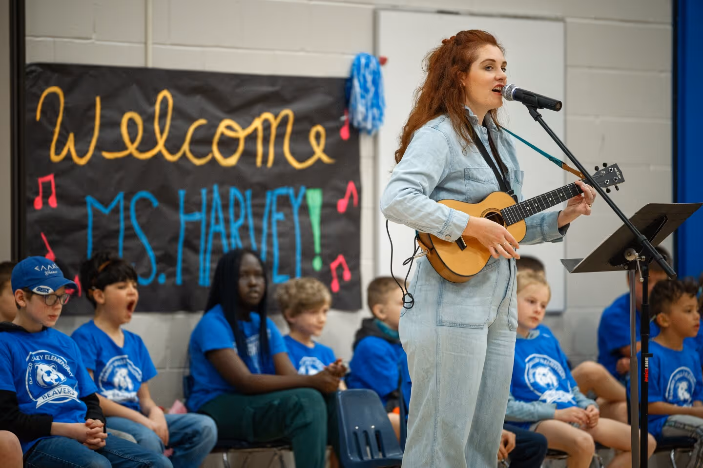 Mandy Harvey sings and plays the ukulele in front of students at Bamber Valley Elementary School. She stands beside a microphone with a sign behind her that reads “Welcome Ms. Harvey!” while children in blue T-shirts watch from their seats.