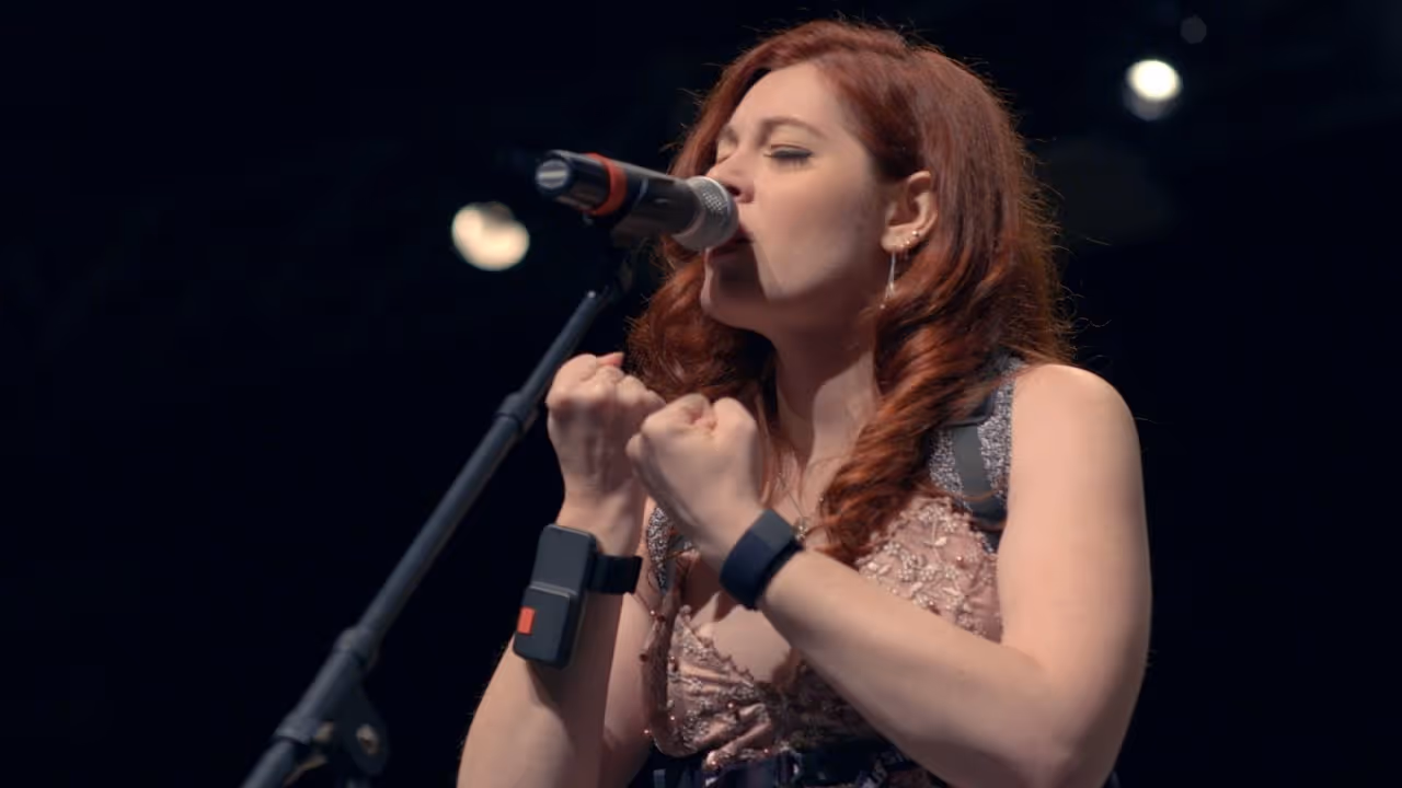 Close-up of Mandy Harvey singing into a microphone, eyes closed with emotion and both fists clenched near her face. She wears a pink embroidered dress and a wristband device while performing under concert lighting.