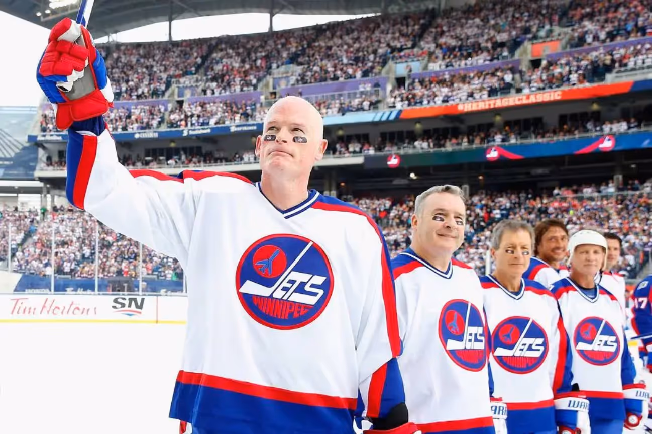 A group of former Winnipeg Jets players stands on the ice in front of a packed stadium during the NHL Heritage Classic. The player in the foreground, wearing a vintage-style Jets jersey and eye black, raises his hockey stick toward the crowd in a gesture of recognition. Teammates behind him smile and look on. The scene conveys celebration and nostalgia.