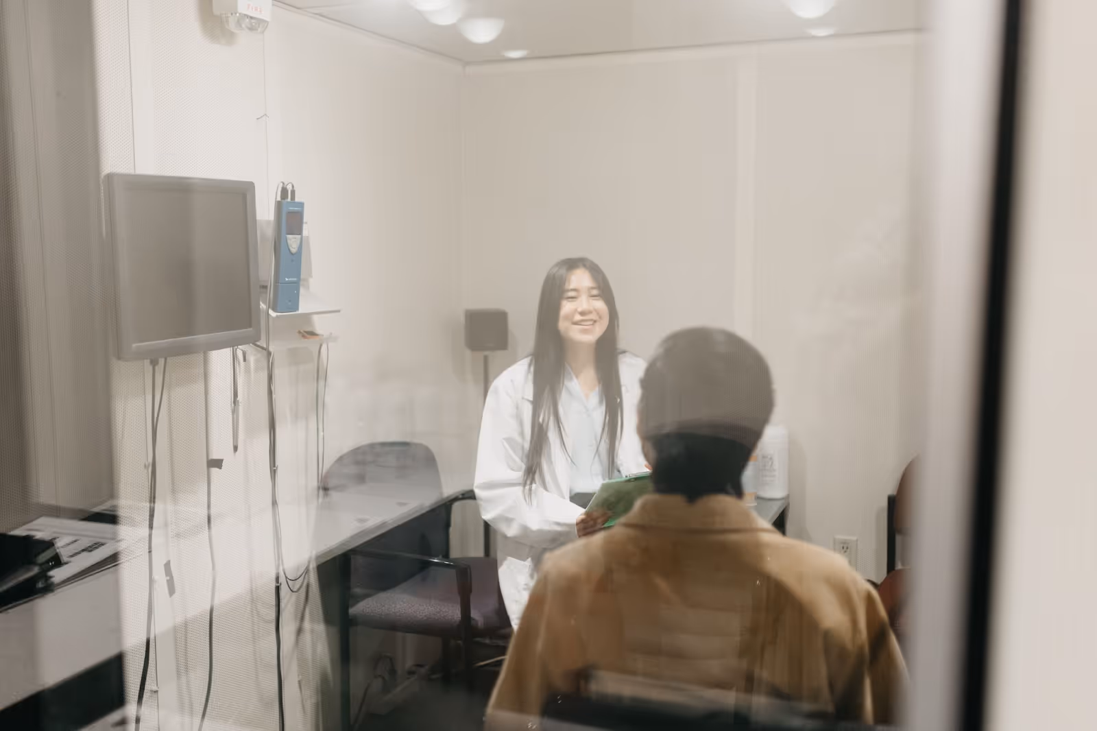 A female audiologist speaks with a patient in a soundproof booth. The image is taken through the booth’s glass window, capturing both participants and hearing test equipment in the room.