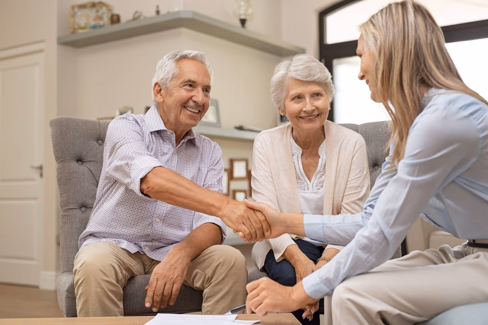 An older couple smiles while shaking hands with a professional woman seated across from them. They appear to be in a warm, home-like setting, possibly during a consultation.
