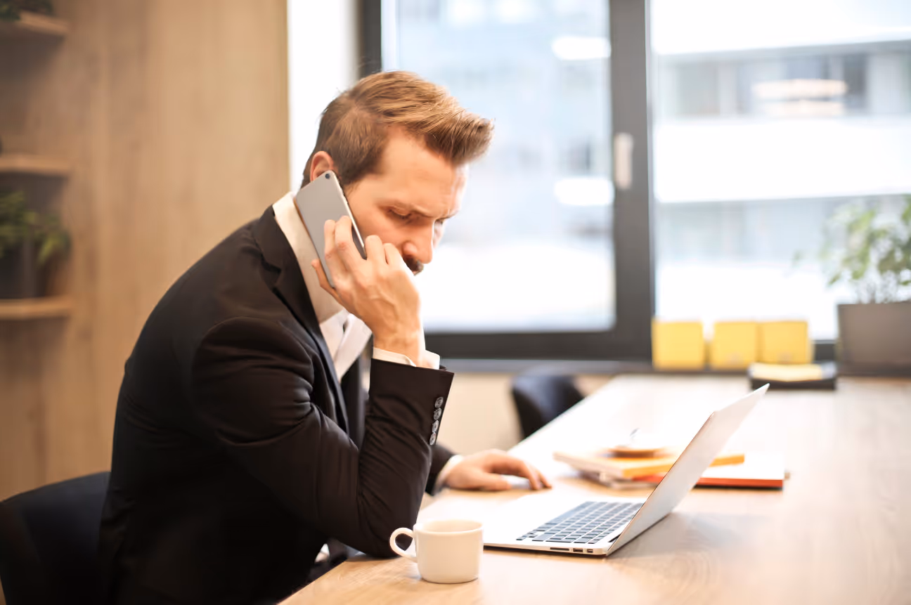 A man in a black suit sits at a desk in a modern office, holding a smartphone to his ear with a serious expression. An open laptop, coffee cup, and notepad are on the table in front of him.