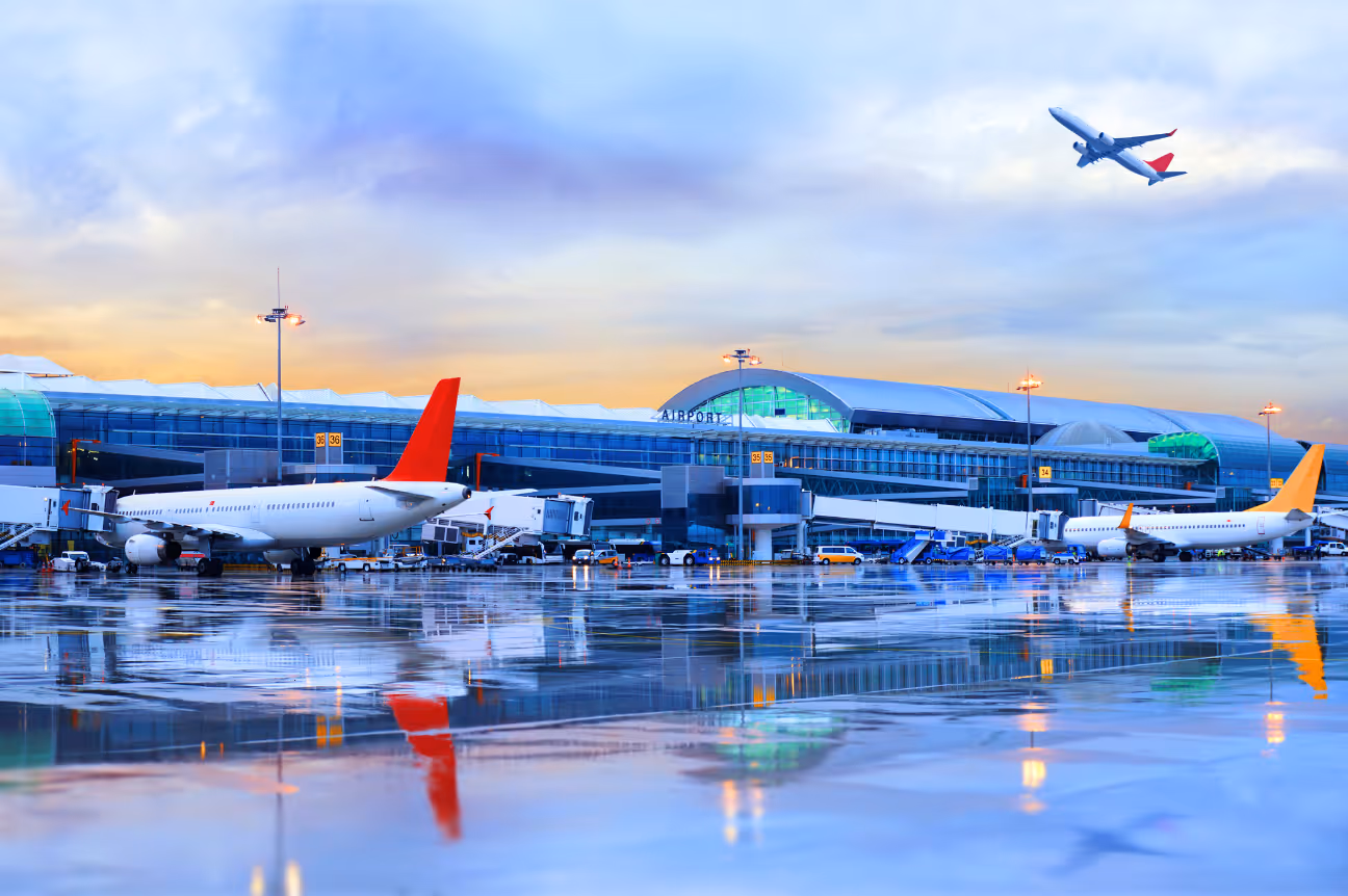 Exterior view of an airport terminal at sunset with planes on the tarmac and one airplane taking off in the colorful sky.